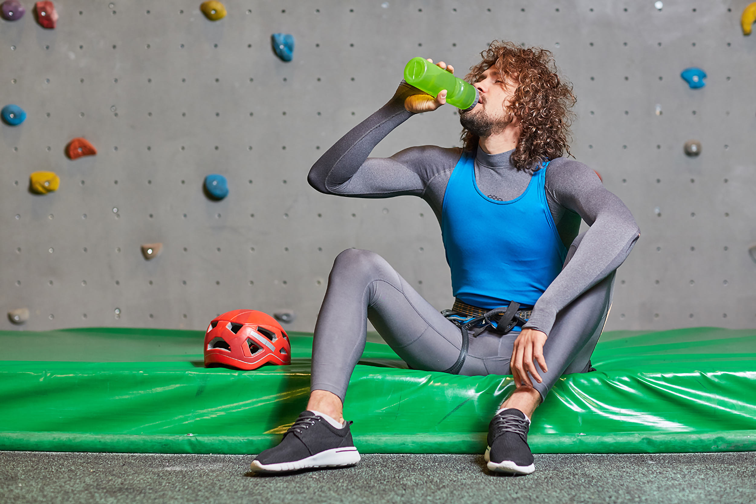 Sporty guy in activewear for climbing sitting on mat and drinking water from plastic bottle after training